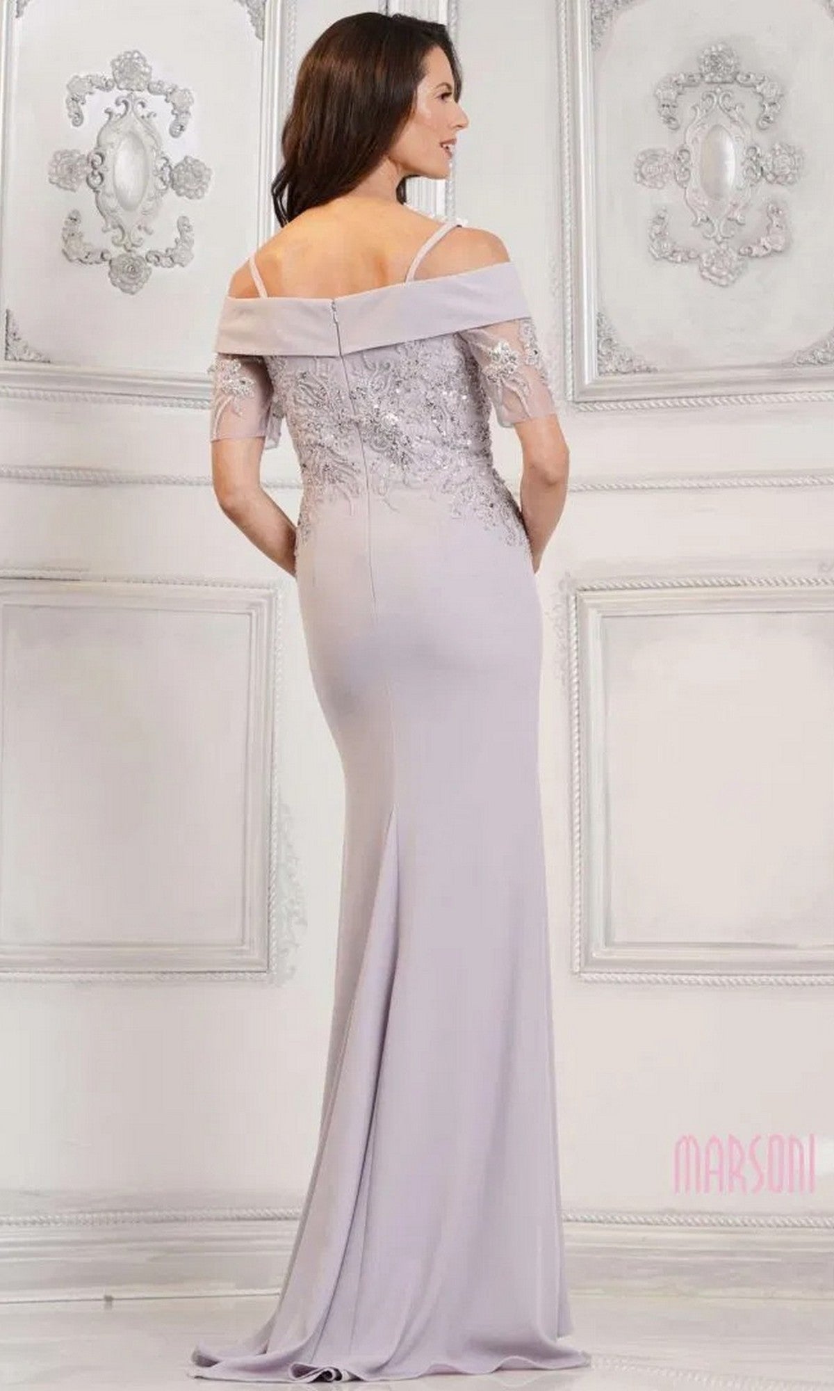 A woman stands in an ornate, light-colored room wearing the Marsoni MV1295 Long Formal Dress, a floor-length lavender gown with off-the-shoulder lace details and sequin-embroidered appliques.